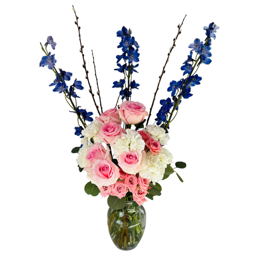 Bouquet of pink, white, and blue flowers in a clear vase on a white surface with a window in the background.